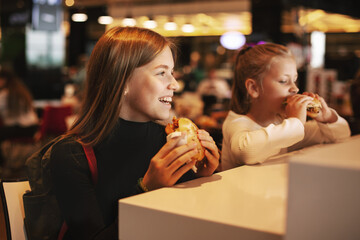 Beautiful schoolgirl eating hamburger sitting in a cafe indoors. Happy child eating junk food in a restaurant. Side view