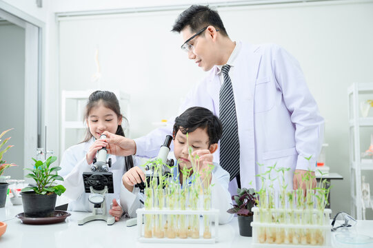 Kids learning about botany hands on. There is a diverse group of children gathered around a microscope and surrounded by plants.