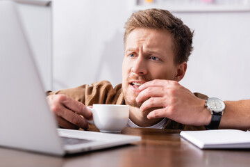 Selective focus of focused businessman with cup of coffee looking at laptop at table