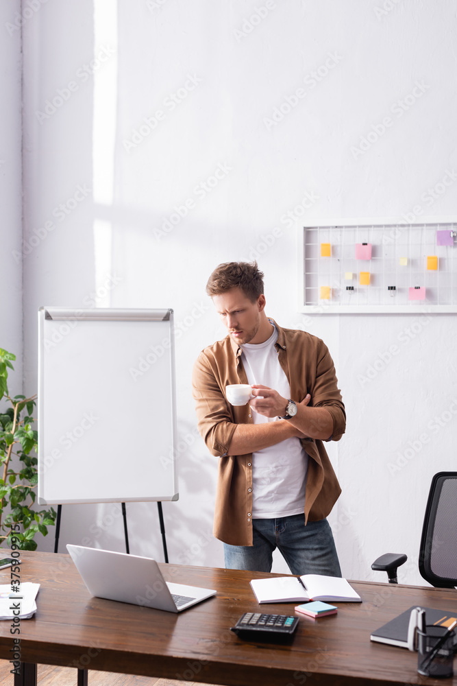 Wall mural selective focus of pensive businessman looking at laptop while holding cup of coffee in office