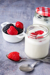 Top view of two spoons, bowl with raspberries, blackberries and two jars of yogurt with raspberries, on gray background, in vertical, with copy space