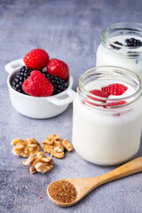 Top view of wooden spoon with brown sugar, walnuts, bowl with raspberries, blackberries and two jars of yogurt with raspberries, on gray background, in vertical, with copy space