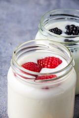 Top closeup of two glass jars with raspberry and blackberry yogurt on defocused gray background, vertical