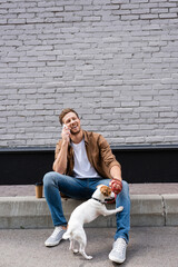 Man holding jack russell terrier on leash while talking on smartphone near coffee to go on urban street