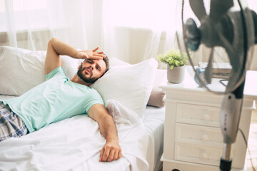 Sweaty young man is trying to refresh from the heat with a fan while lying in bed at home.