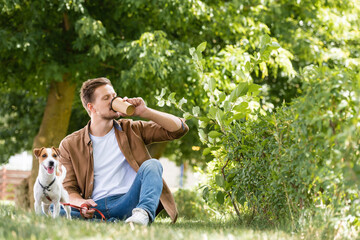 man in casual clothes drinking coffee to go while sitting on lawn near jack russell terrier dog
