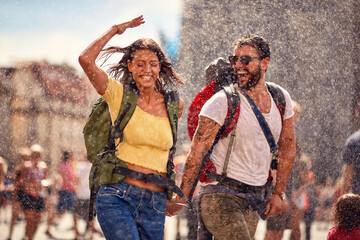 Young cheerful tourist couple enjoying the fountain shower at a city square