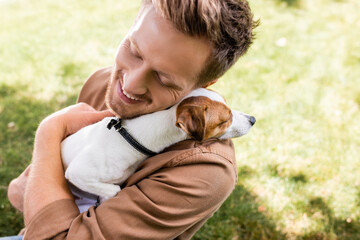 high angle view of young man with closed eyes holding white jack russell terrier dog with brown...