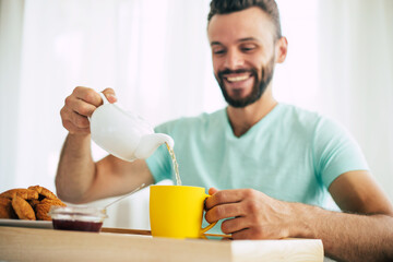 Happy young bearded man is sitting on his bed and has his breakfast with tea or coffee