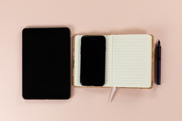 View of a tablet and phone with notebook and pen on pink background
