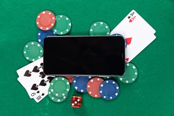 View of a black smartphone, playing cards, a red dice and colorful tokens on plain green surface
