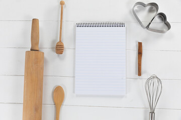 View of a composition with a notebook with cookie cutters, whisker and rolling pin on a white wooden