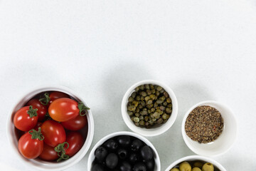 View of a six bowls with fresh tomatoes, olives, nuts and seasoning on plain white surface
