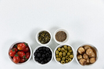 View of a six bowls with fresh tomatoes, olives, nuts and seasoning on plain white surface