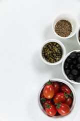 View of a six bowls with fresh tomatoes, olives, nuts and seasoning on plain white surface