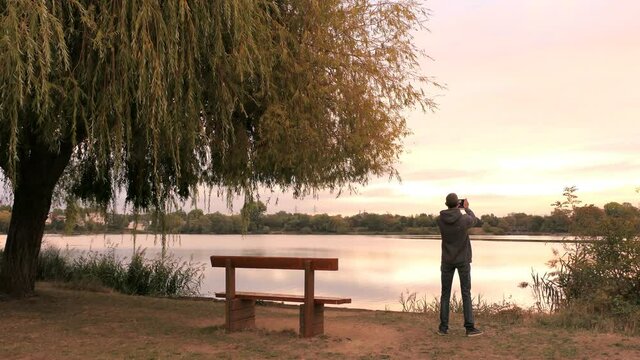 Man photographing with his smartphone. Lake and weeping willow tree at sunrise or sunset. Romantic landscape with a lonely bench.	