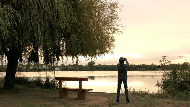 Man photographing with his smartphone. Lake and weeping willow tree at sunrise or sunset. Romantic landscape with a lonely bench.	