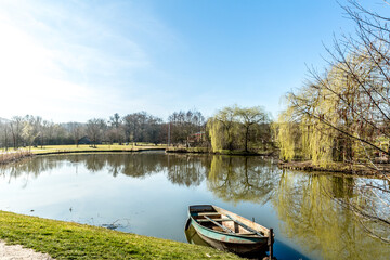 Obraz premium Lake in the middle of a park with beautiful reflection in the water and a small boat anchored on the shore, surrounded by trees on a wonderful day in Landgraaf in the Netherlands