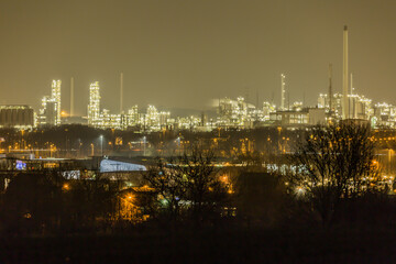 Fototapeta na wymiar Industrial complex with its metal structures illuminated by white lights seen from a hill with bare trees, quiet night in southern Limburg in the Netherlands Holland