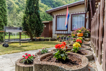 Fototapeta na wymiar Stone pots with red, yellow and pink flowers on the porch of a country house with a Dutch flag between two windows, a small garden with green grass and trees, sunny and relaxing day in Schuld, Germany