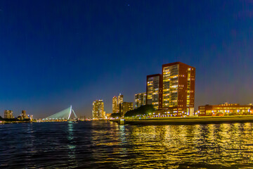 Obraz premium Night cityscape in Rotterdam with its buildings with lights on and the Erasmus cable-stayed bridge over the Maas river, clear blue sky in the Netherlands
