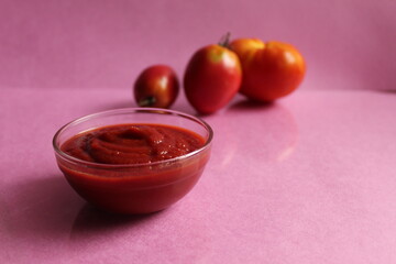 red tomato sauce in a glass saucepan plate next to the back are vegetables tomatoes and onions on a pink background copy space