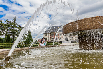 Woman wearing white casual clothes sitting on the shore of a fountain, seen through the jets of water, sunny day in a villa with a blue sky and many white clouds