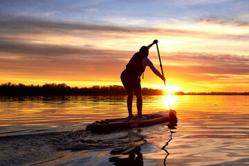 Silhouette of woman paddle on stand up paddle boarding (SUP) on quiet winter or autumn river at sunset. Colorful sunset over the river