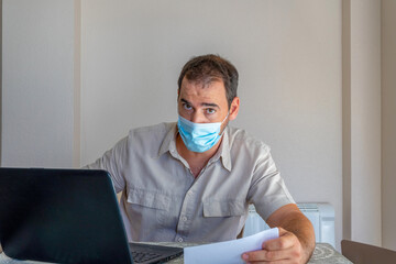 Fototapeta na wymiar Bearded man dressed in beige shirt and wearing a mask to protect himself from coronavirus looking at bills on computer worried in his living room at home