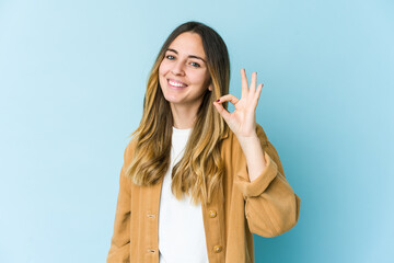 Young caucasian woman isolated on blue background cheerful and confident showing ok gesture.