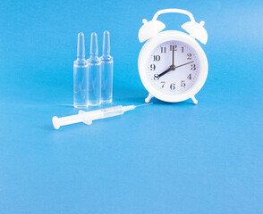 White alarm clock, medical ampoules, syringe on a blue background with copy space. The concept of vaccination against infection and viruses