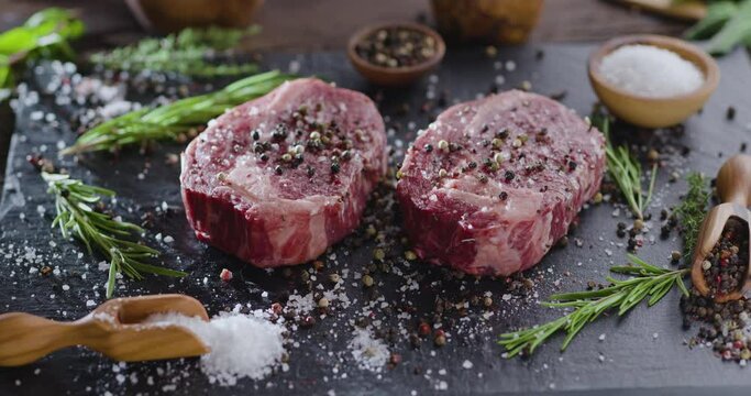 Two raw rib eye steaks with spices and herbs on a stone surface. The camera rotates slowly around the steaks.