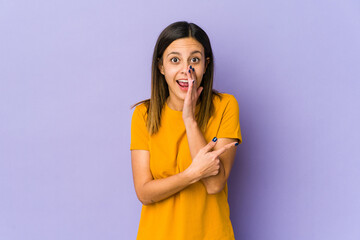 Young woman isolated on purple background saying a gossip, pointing to side reporting something.