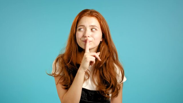 Beautiful woman in overall holding finger on her lips over blue background. Gesture of shhh, secret, silence. Close up.