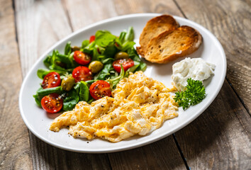 Delicious breakfast - scrambled eggs with fried sausages, cottage cheese and vegetables served on black plate on wooden table

