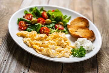 Delicious breakfast - scrambled eggs with fried sausages, cottage cheese and vegetables served on black plate on wooden table
