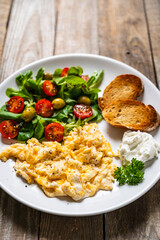 Delicious breakfast - scrambled eggs with fried sausages, cottage cheese and vegetables served on black plate on wooden table
