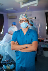 Doctor stands cross hands. Blue scrubs. Robotic medical equipment background. Medical team working. Doctor posing.