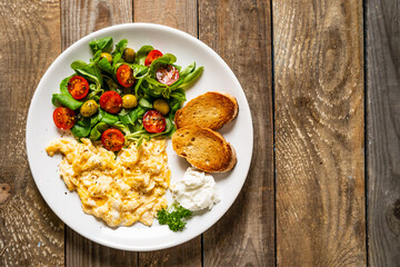 Delicious breakfast - scrambled eggs with fried sausages, cottage cheese and vegetables served on black plate on wooden table
