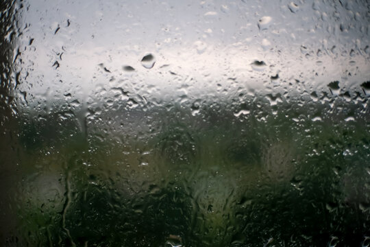Raindrops on the window on a rainy gloomy day. Melancholy view through the wet glass of the gray sky, green dark trees outside. The concept of sadness, depression, and sadness