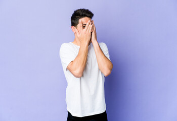Young caucasian man isolated on purple background blink through fingers frightened and nervous.