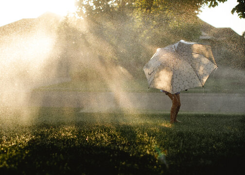 Child playing in the sprinklers holding an umbrella at sunset