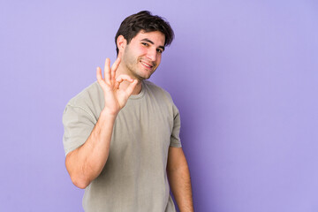 Young man isolated on purple background winks an eye and holds an okay gesture with hand.