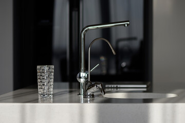 Glass of pure filtered water on the kitchen table in the rays of sunlight. Kitchen faucet and reverse osmosis faucet for water purification. Healthy lifestyle concept. Сlose-up shot, selective focus.