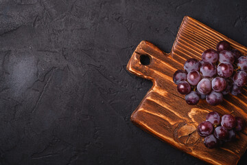 Fresh ripe grape berries on brown wooden cutting board on dark stone background, top view copy space
