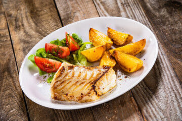 Fish dish - fried cod fillet with potatoes and vegetable salad on wooden table