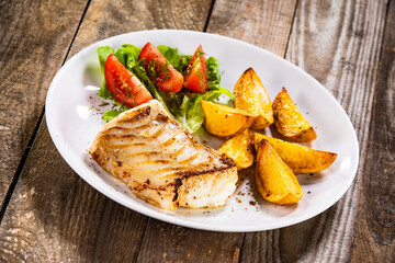 Fish dish - fried cod fillet with potatoes and vegetable salad on wooden table