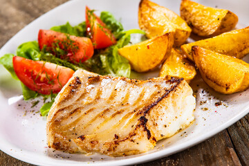 Fish dish - fried cod fillet with potatoes and vegetable salad on wooden table