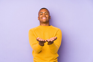 Young latin man isolated on purple background holding something with palms, offering to camera.