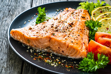 Fried salmon fillet with lettuce, lemon and tomatoes served on black plate on wooden table
